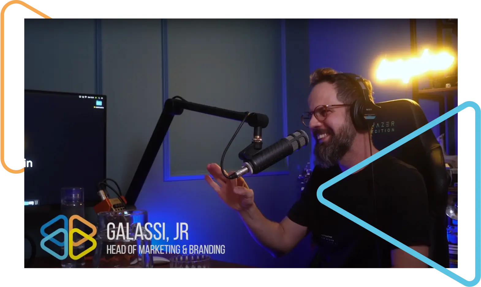 Homem branco, de óculos, cabelo castanho, bigode e barba, sentado em um estúdio de gravação de áudio